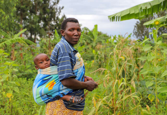 En mamma går genom grönskande fält med ett litet barn på ryggen.