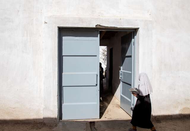 Eine Frau in einem schwarzen Gewand geht durch eine halbgeöffnete Tür in einer weißen Wand.