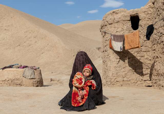 A woman, wrapped in a blanket, holds her young grandson in her arms outside of a clay structure in rural Afghanistan.