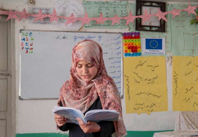 A female student reads from a book while standing at the front of a classroom in Afghanistan.