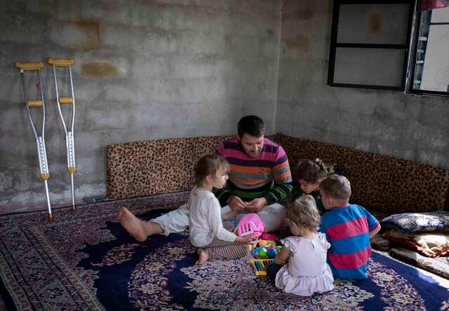 Eine Mann sitzt mit vier kleinen Kindern in einem möblierten Raum auf dem Boden. Sie sitzen alle auf einem großen Teppich.