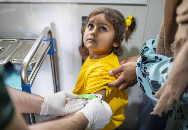 Ein kleines Mädchen in einem gelben T-Shirt erhält eine Impfung in den Arm, während eine Person sie sanft festhält.