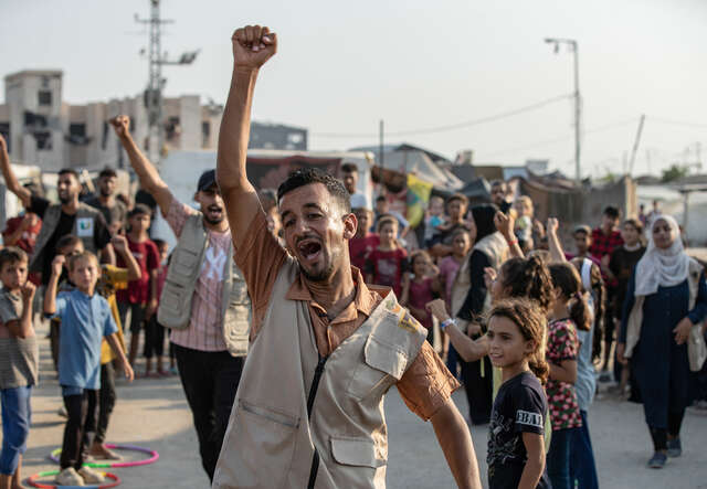 Nour holds his hand up in the air, with children cheering around him.