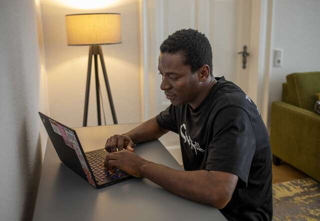 Ein Mann sitzt an einem Schreibtisch und arbeitet an einem Laptop in einem Raum mit einer Stehlampe und einem grünen Sofa im Hintergrund.