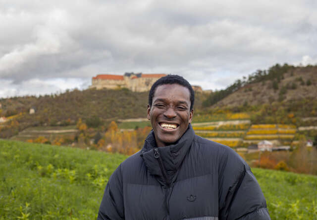 Eine lachende Person steht vor grünen Weinbergen mit einem Schloss auf einem Hügel im Hintergrund unter einem bewölkten Himmel.