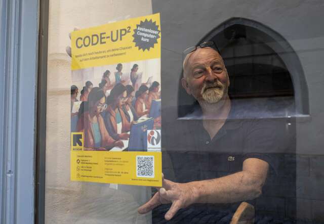 Ein lächelnder Mann steht hinter einer Glastür, neben der ein gelbes Plakat mit Menschen und Text angebracht ist.