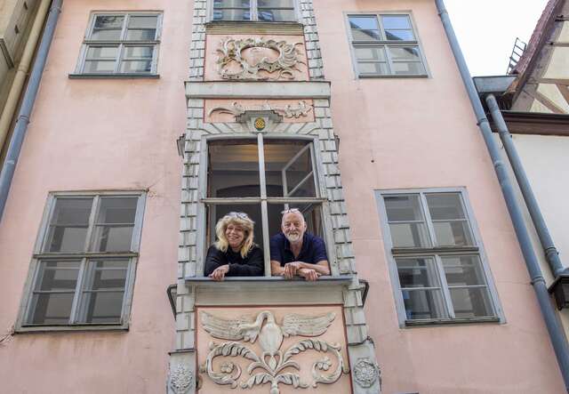 Zwei Personen stehen an einem geschmückten Balkon eines historischen Gebäudes mit bemalter Fassade und mehreren Fenstern.