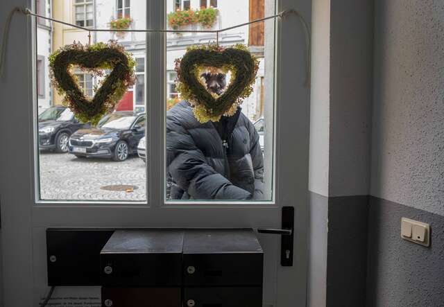 Eine Person steht draußen und blickt durch eine verglaste Tür mit zwei herzförmigen Dekorationen, während im Hintergrund Autos zu sehen sind.