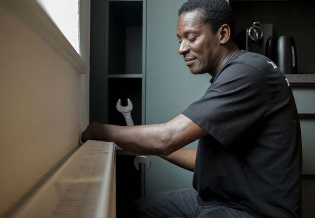 Ein lachender Mann sitzt an einem Tisch in einem Innenraum und arbeitet konzentriert mit seinen Händen an einem Gegenstand.