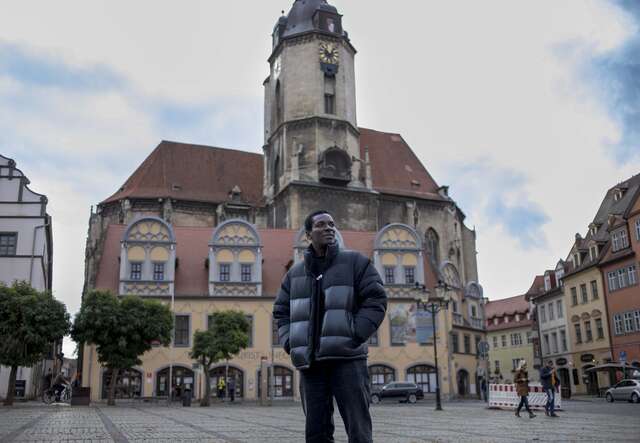 Eine Person steht vor einem historischen Gebäude mit Turm, verziertem Dach und mehreren Fenstern auf einem Platz unter einem leicht bewölkten Himmel.