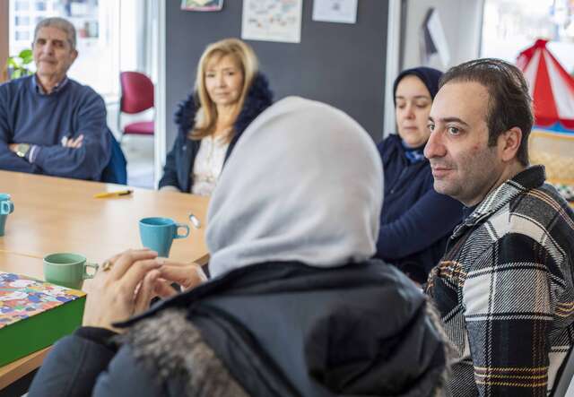 Eine Gruppe von Menschen sitzt an einem Tisch in einer lockeren Gesprächsrunde, während eine Person mit Kopftuch im Vordergrund zu sehen ist.