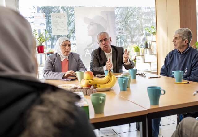Eine Gruppe von Menschen sitzt an einem Tisch mit Obst und Tassen, während eine Person im Vordergrund mit dem Rücken zur Kamera sitzt.