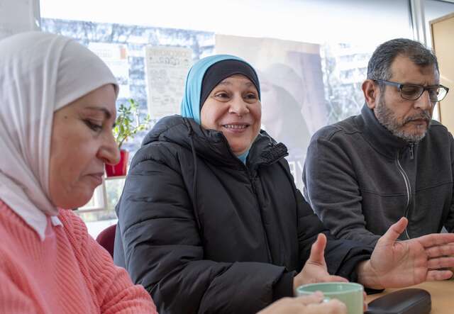 Drei Personen sitzen an einem Tisch, zwei Frauen mit Kopftüchern und ein Mann, während eine der Frauen lächelnd in die Kamera schaut.