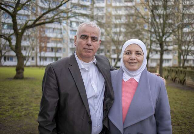 Ein Mann und eine Frau mit Kopftuch stehen zusammen in einem Park, mit Wohngebäuden im Hintergrund, und blicken in die Kamera.