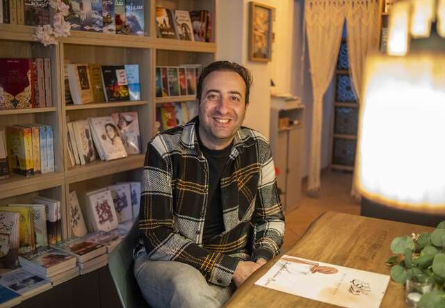 Ein Mann sitzt an einem Tisch in einem Raum mit Bücherregalen im Hintergrund und lächelt in die Kamera.