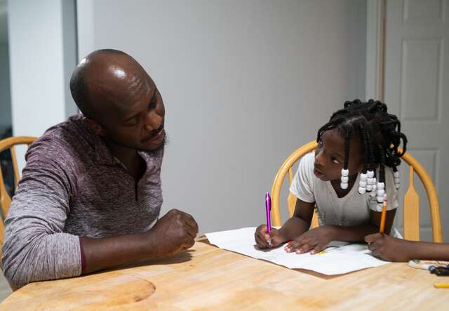 A father helps his daughter with a homework assignment in their living room.