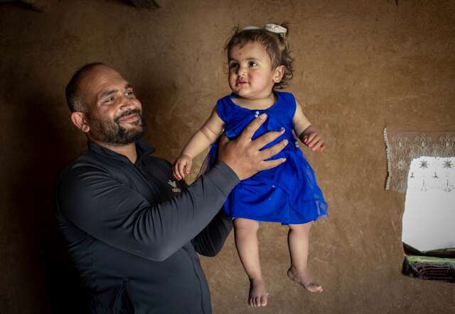 A father holds his youngest daughter in the air in their home in Syria. Both share a smile.