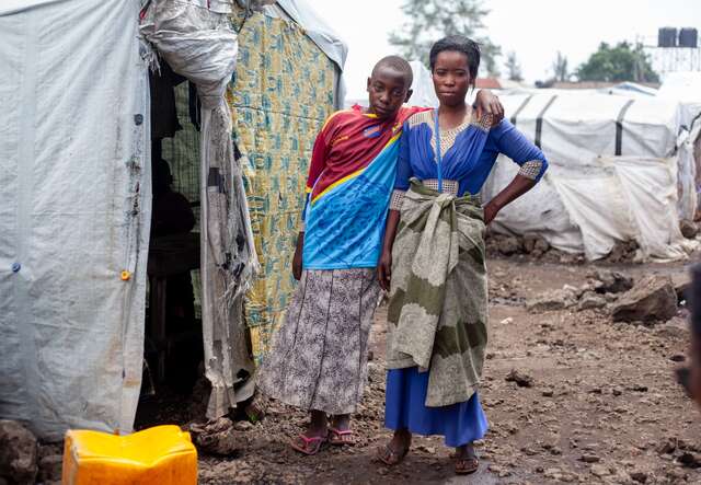A son stands next to his mother outside of their shelter in a camp for displaced people in the DRC.