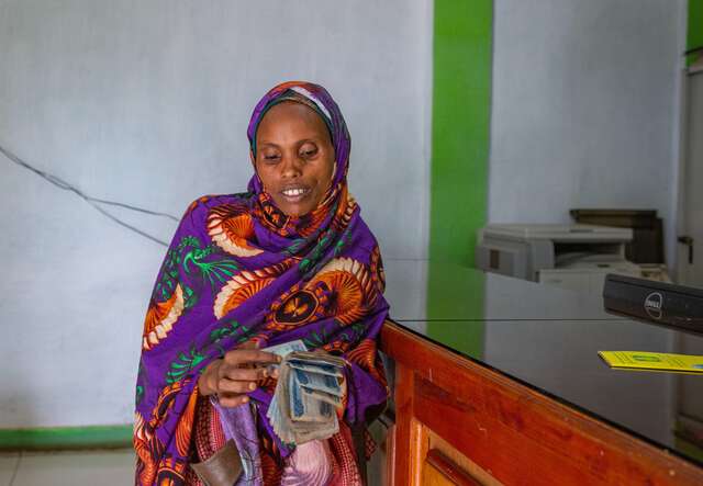 Junge Frau in farbenfroher Kleidung zählt lächelnd Geld in einem einfachen Raum.