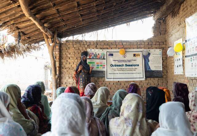 Eine Frau hält eine Aufklärungssitzung vor einer Gruppe von Frauen in einem einfachen, überdachten Raum.