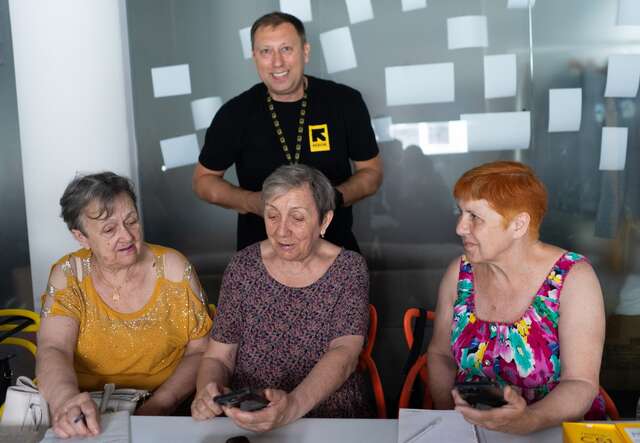Drei Frauen sitzen an einem Tisch und unterhalten sich, während ein Mann im Hintergrund steht und lächelt.