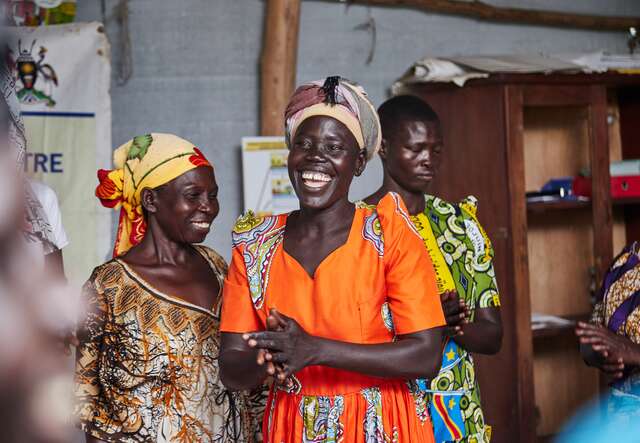 Drei Frauen in bunter traditioneller Kleidung stehen lachend in einem Raum und wirken fröhlich und lebendig.