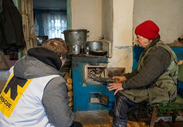 A woman wearing a red hat putting firewood into the stove with an IRC staff member watching her
