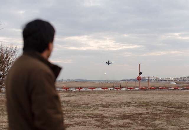 A man looks away in the distance at a plane that is taking off from an airport.