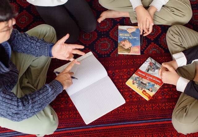 An overhead shot of a man counting on his fingers and teaching lessons to his children who sit around him.