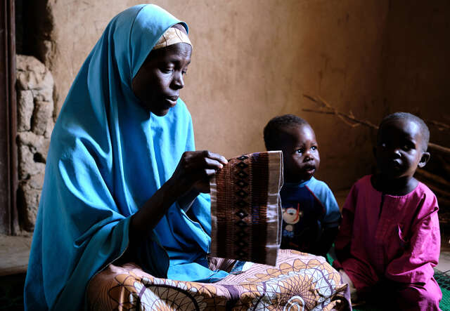 En mamma sitter på golvet och syr en traditionell nigeriansk hatt. Bredvid henne sitter hennes två söner.