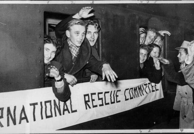 Svartvid bild på unga män och kvinnor som sticker ut sina huvuden från tågfönster, med en banderoll där det står International Rescue Committee.