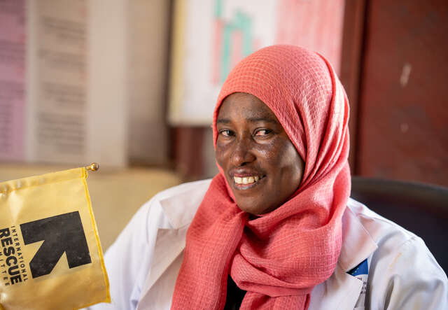 En kvinna i läkarrock ler mot kameran, bredvid henne syns RESCUE:s logga.