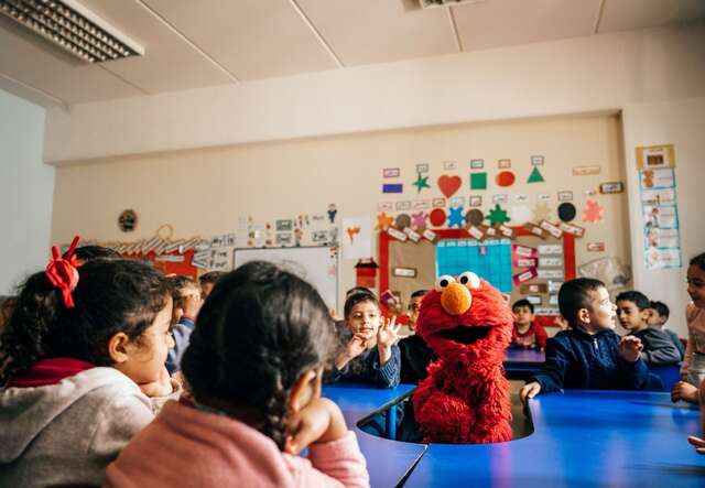 Barn i ett klassrum sitter runt en färgglad karaktär från Sesame street.
