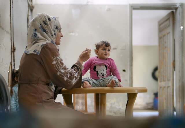 A woman wearing a hijab feeding her young daughter who sits on a table in a pink sweater with Minnie Mouse