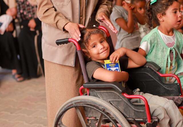 A young Palestinian girl in a wheelchair holds a pack of coloured pencils in her arms and smiles.