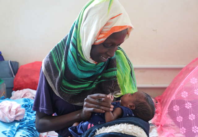 한 엄마가 아기의 손을 잡고 미소 지으며 아기를 바라보고 있습니다.