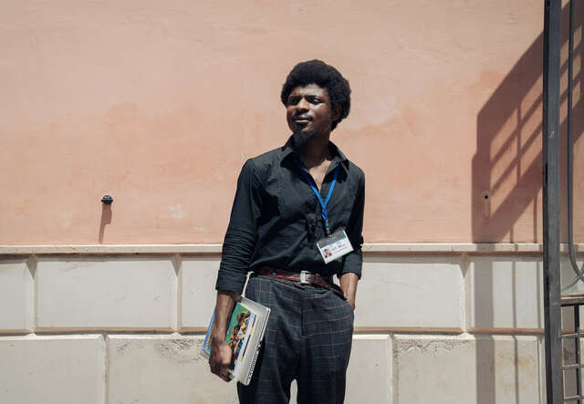 Abubakar stands in front of a wall, holding some books.