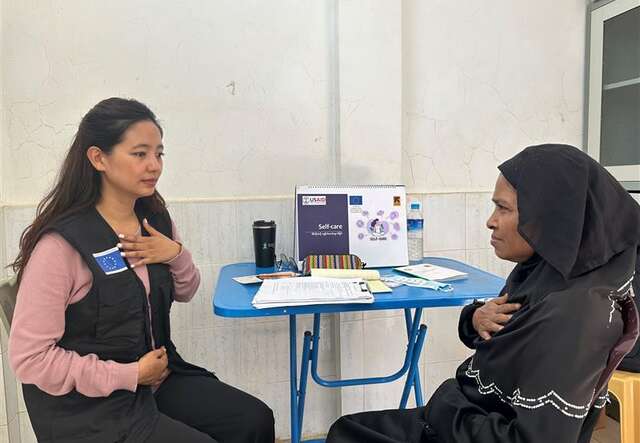 A young woman with long brown hair and an IRC/EU ECHO vest is talking to another woman across the table from her. The second woman is wearing a brown dress and head scarf. 