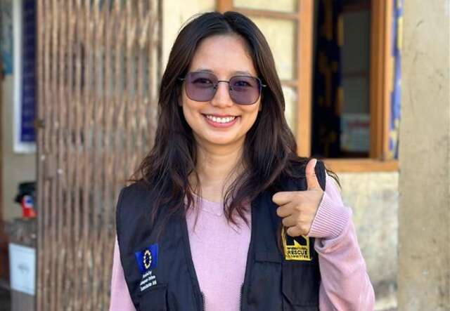 A young woman with long brown hair and sunglasses is looking at the camera and doing a thumbs up with her left hand