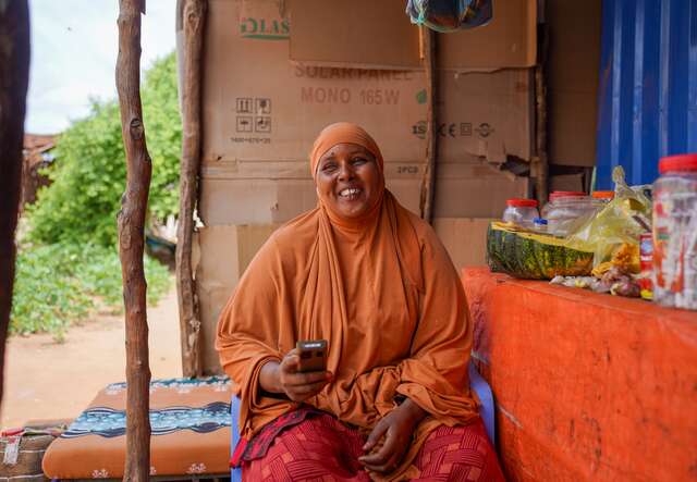 Somalische Frau in orangefarbener Kleidung sitzt mit Handy in der Hand vor einem kleinen Verkaufsstand.