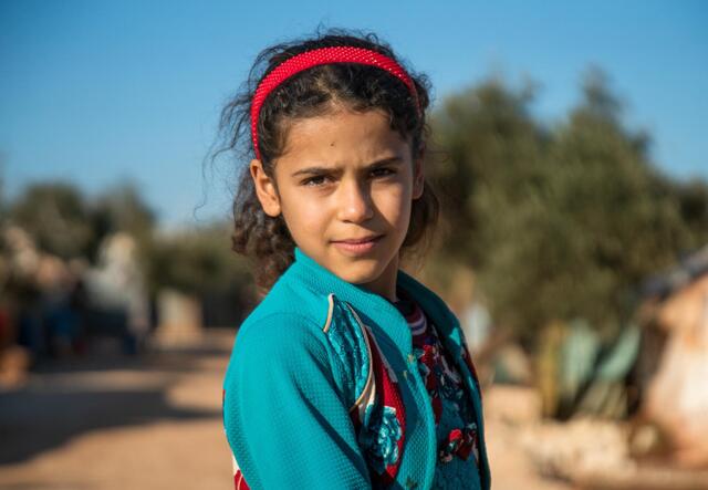 10-year-old Heba looks at the camera. She wears a red head band and green cardigan.