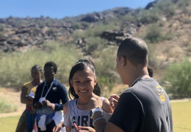 In the forefront, a refugee girl laughs with an IRC staff member. In the background three girls stand.