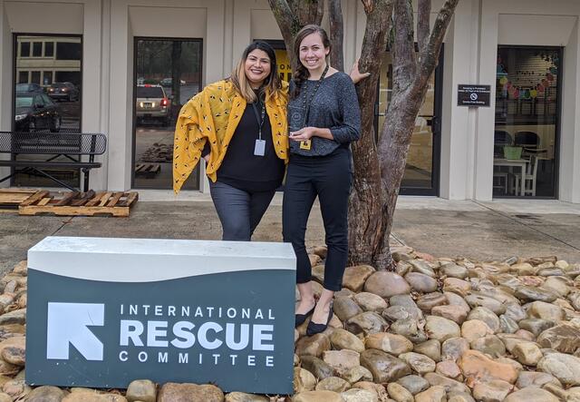 Two women stand beside a large sign that says International Rescue Committee and just in front of a medium sized tree. They are smiling and holding glass awards. They wear lanyards and work badges around their necks.