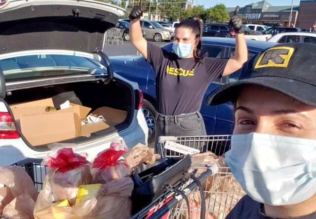 Two IRC staff members wearing IRC shirts and hats stand next to a car with an open trunk. In the trunk are groceries they recently bought for refugee and immigrant families.