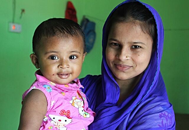 Lovely Akter, an IRC midwife, poses with her nine-month-old daughter. Lovely is wearing a blue headscarf and her daughter is wearing a pink shirt. Both are smiling.
