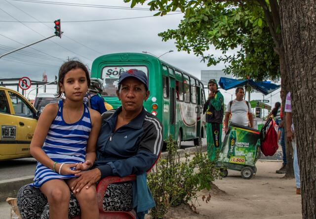 Venezuelan mother Karina sits with her ten-year-old daughter Geicelis on her lap. They are sitting outside looking at the camera. There is a green bus behind them.