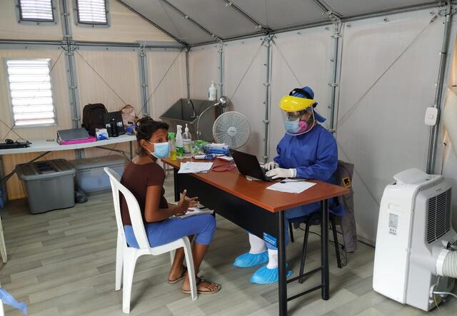 A woman sits at a chair at a table with doctor in full PPE opposite her. They are in a temporary clinic set up by the IRC at the Simón Bolívar bridge.