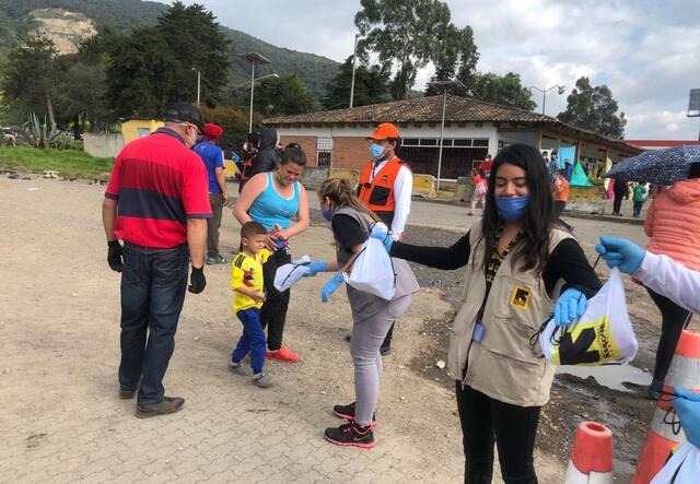 Two IRC staff members stand in a line passing out supplies outside near the Bogota-Chia highway. A family, including a young boy, are given supplies.