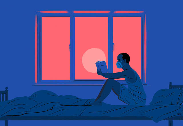 A man sits on a bed reading a book in front of a window.