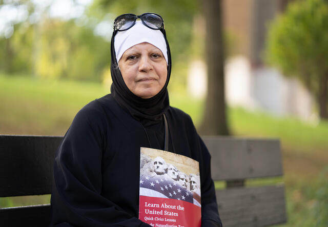 Maha, a refugee from Iraq and new American citizen, sits on a park bench in Queens, NY, holding a book on the U.S. citizenship test.